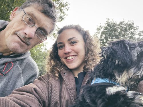 ein Mann mit Brille und eine junge Frau mit Locken schauen in die Kamera und machen ein Selfie; die Frau trägt einen kleinen Hund im Arm