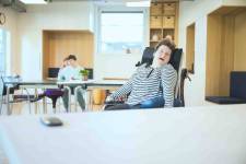 Eine Frau im Rollstuhl sitzt am Tisch. Im Hintergrund zwei Personen.