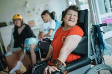 Eine Frau mit rotem Hemd sitzt im Rollstuhl und lächelt. Im Hintergrund zwei weitere Frauen. Eine davon sitzt im Rollstuhl.