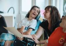 Eine Frau im Rollstuhl mit weißem Hemd lächelt eine Unterstützerin an. Die Unterstützerin hält ein Blatt Papier.