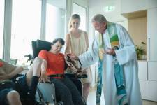 Ein Kapuzinerbruder in weißer Kuttel berührt eine Frau im Rollstuhl auf den Arm. 