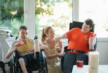 Ein Mann im Rollstuhl und eine Frau im Rollstuhl tauschen sich aus mit einer Unterstützerin, die zwischen denen sitzt.