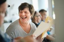 Eine Frau mit gestreiftem Hemd schaut lächelnd nach rechts und hält ein Blatt Papier.