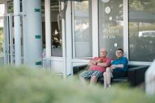 Ein Mitarbeiter des Pastoralteams und ein Klient mit blauem Hemd sitzen nebeneinander auf einer schwarzen Couch im Freien. Im Vordergrund ist grünes Gebüsch zu sehen. Im Hintergrund sind große Fensterflächen. 