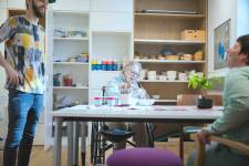 Eine Klientin des slw sitzt alleine am Tisch und bemalt eine Keramikschüssel. Ein Unterstützer steht daneben und unterhält sich. Im Hintergrund sind Regale zu sehen, auf denen mehrere Keramikschüssel, Keramiktassen und Keramikteller gestapelt sind.