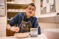 Ein Klient mit Brille sitzt am Tisch und bemalt eine Keramikschüssel. Vor ihm ein großer Farbtegel. In der rechten Hand hält er der Pinsel. Er lächelt.