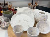 Drei weiße Keramiktassen und ein Teller mit bunte Farbtupfen stehen auf ein Tisch, sowie mehrere Becher in denen Pinsel stecken.