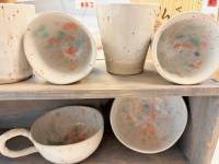 Eine helle Keramiktassen, ein Schüssel und drei helle Keramikbecher stehen übereinander auf zwei Regale. 