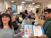 Eine Mitarbeiterin und ein Klient des slw stehen im Vordergrund und halten gemeinsam ein Buch mit  Piktogrammen aus der Unterstützten Kommunikation. Im Hintergrund sitzen mehrere Klienten und Klientinnen sowie Mitarbeitende des slw gemeinsam am Tisch.