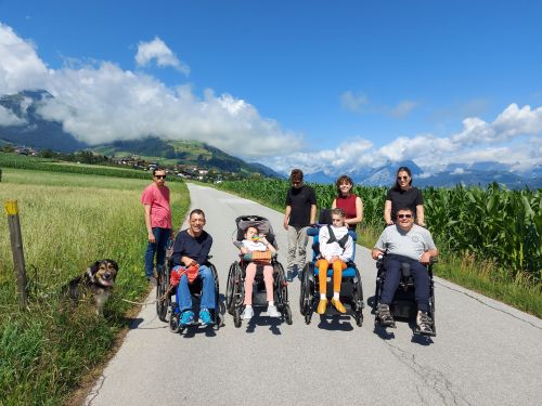 eine Gruppe Menschen mit und ohne Behinderungen spazieren auf einem Feldweg an einem schönen, sonnigen Tag; Manche Menschen sitzen im Rollstuhl; links im Gras sitzt ein schwarz-brauner Hund; alle schauen freundlich in die Kamera