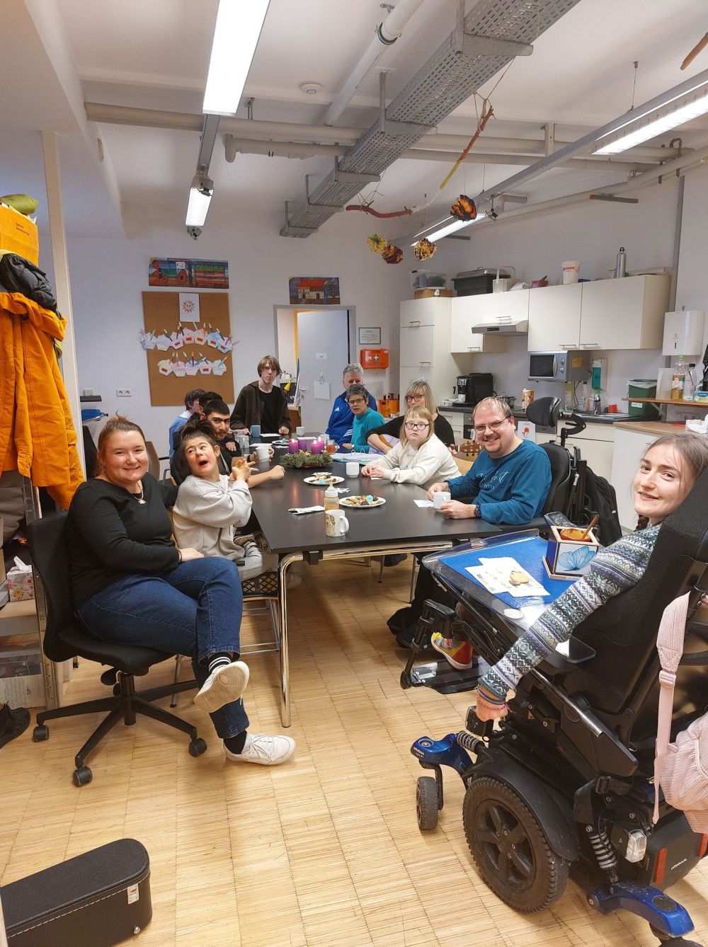 eine Gruppe Menschen mit und ohne Behinderungen sitzen um einen Tisch herum und lächeln in die Kamera