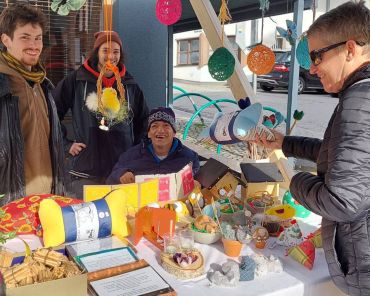 drei Männer bieten Waren an einem Marktstand an; eine Kundin mit Sonnenbrille sieht sich die Waren an