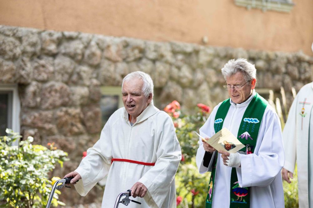 ein älterer Mann mit Behinderung geht in einer Kutte eines Ministranten; neben ihm ein Priester; er hält einen Folder in der Hand