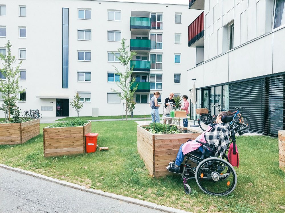 ein Garten in einer Hochhausanlage; mehrere Hochbeete; eine Person sitzt im Rollstuhl, einige weitere Personen stehen um die Hochbeete