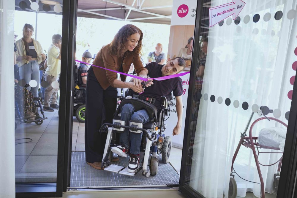 eine Frau und ein Mann im Rollstuhl schneiden gemeinsam mit einer Schere ein pinkes Band durch; das Band ist in einem Türrahmen gespannt