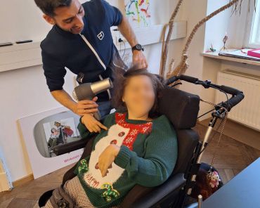 Eine Frau im Rollstuhl hält einen Föhn an ihre Haare, ein Unterstützer steht daneben und hilft ihr