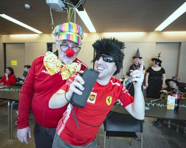ein Mann ist als Clown verkleidet und ein anderer trägt Perücke, Sonnenbrille und Fußballtrikot; sie hören Musik