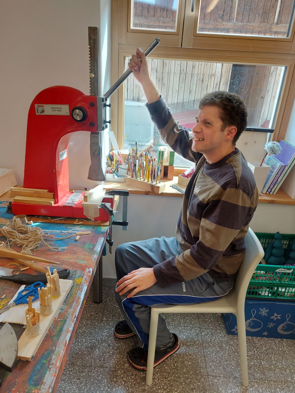 ein Mann sitzt an einer Werkbank und bedient eine rote Presse mit einem langen Hebel