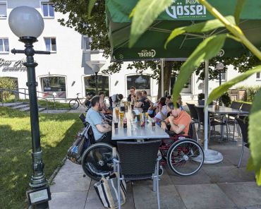 eine Gruppe Erwachsener mit Behinderungen sitzen an einem Tisch in einem Gastgarten; es stehen Getränke und Speisen am Tisch