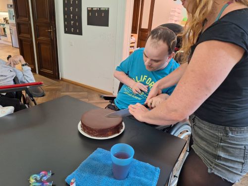 ein Mann mit Behinderung schneidet mit einer Unterstützungsperson einen Schokokuchen an