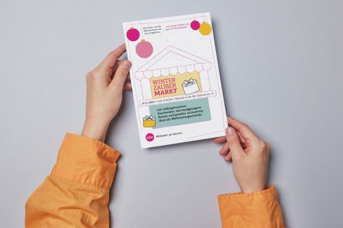 zwei Hände halten einen Flyer; auf dem Flyer ist eine Einladung zum Winterzaubermarkt am 27.11.2024