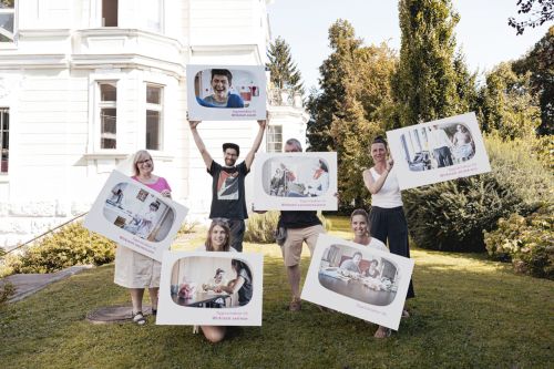 sechs Erwachsene stehen auf einer Wiese und halten Tafeln mit Bildern in die Luft