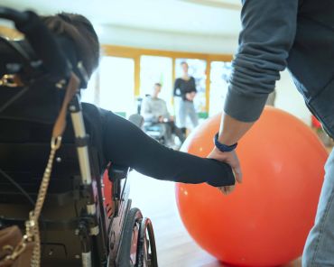 ein Mensch mit Behinderung und ein weiterer halten Hand; man sieht einen großen roten Gymnastikball
