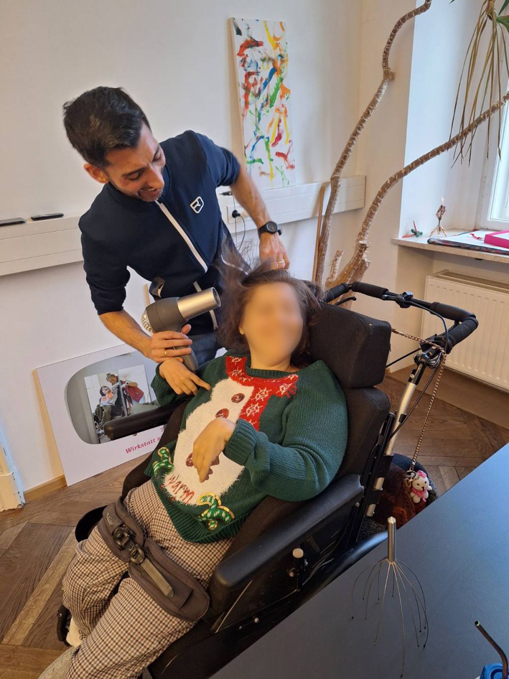 Eine Frau im Rollstuhl hält einen Föhn an ihre Haare, ein Unterstützer steht daneben und hilft ihr
