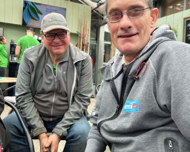 zwei Männer sitzen nebeneinander; im Hintergrund ein Glasdach und Gärtnerei-Mitarbeiter mit grünen Shirts