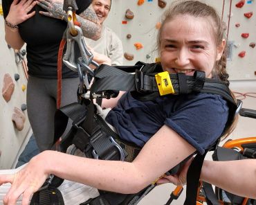 eine Frau mit Behinderung hängt in einem Klettergurt in einer Indoor-Kletterhalle und lächelt in die Kamera