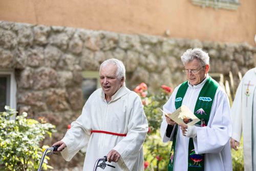 ein älterer Mann mit Behinderung geht in einer Kutte eines Ministranten; neben ihm ein Priester; er hält einen Folder in der Hand