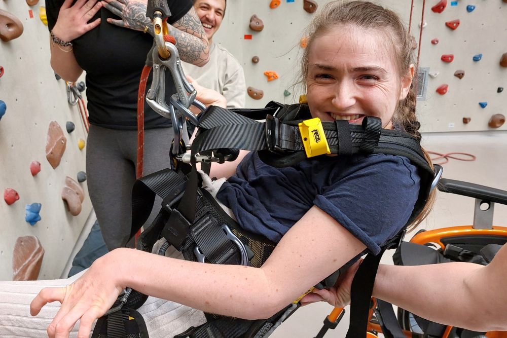eine Frau mit Behinderung hängt in einem Klettergurt in einer Indoor-Kletterhalle und lächelt in die Kamera