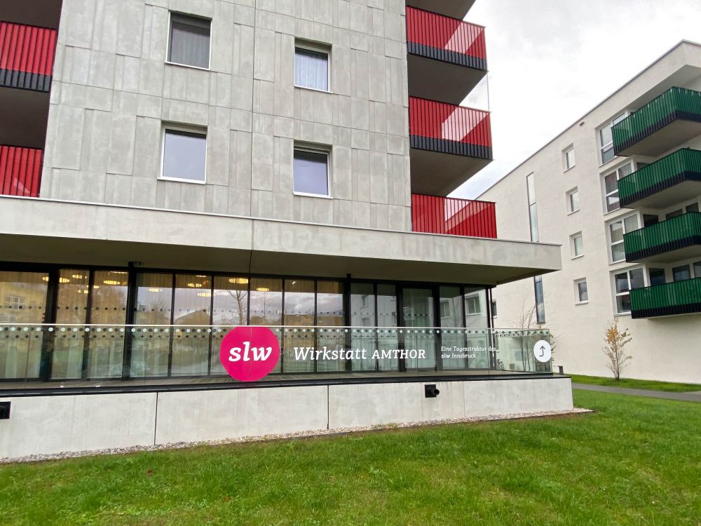 ein Hochhaus mit Grünfläche davor; auf der Glasfläche der Terrassenbegrenzung steht slw Wirkstatt Amthor