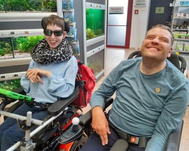 eine Frau und ein Mann mit Behinderung im Rollstuhl in einem Geschäft; beide lächeln in die Kamera