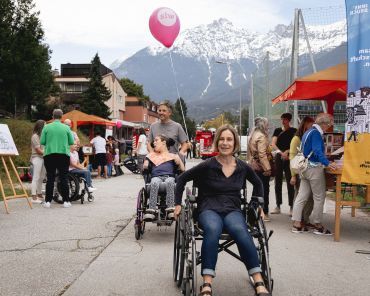eine Frau im Rollstuhl lächelt in die Kamera; dahinter schiebt ein Mann eine weitere Frau im Rollstuhl; pinke Luftballons und bunte Straßenfest-Buden im Hintergrund