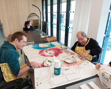 zwei Männer sitzen an einem großen Tisch gegenüber; mit Farbrollen rollen sie weiße Farbe auf kleine Holzstücke