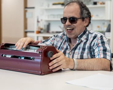 Ein Mann mit Sehbehinderung sitzt an einem Tisch und bedient eine Braille-Schreibmaschine. Er trägt eine Sonnenbrille und lächelt.