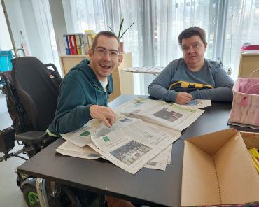 zwei Menschen mit Behinderungen sitzen an einem Tisch und lesen eine Zeitung