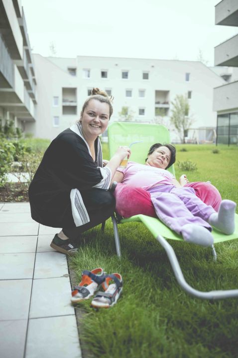 Eine Frau mit Behinderung liegt in einem Liegestuhl in einem Garten. Eine Betreuerin hält ihre Hand. Beide lächeln.