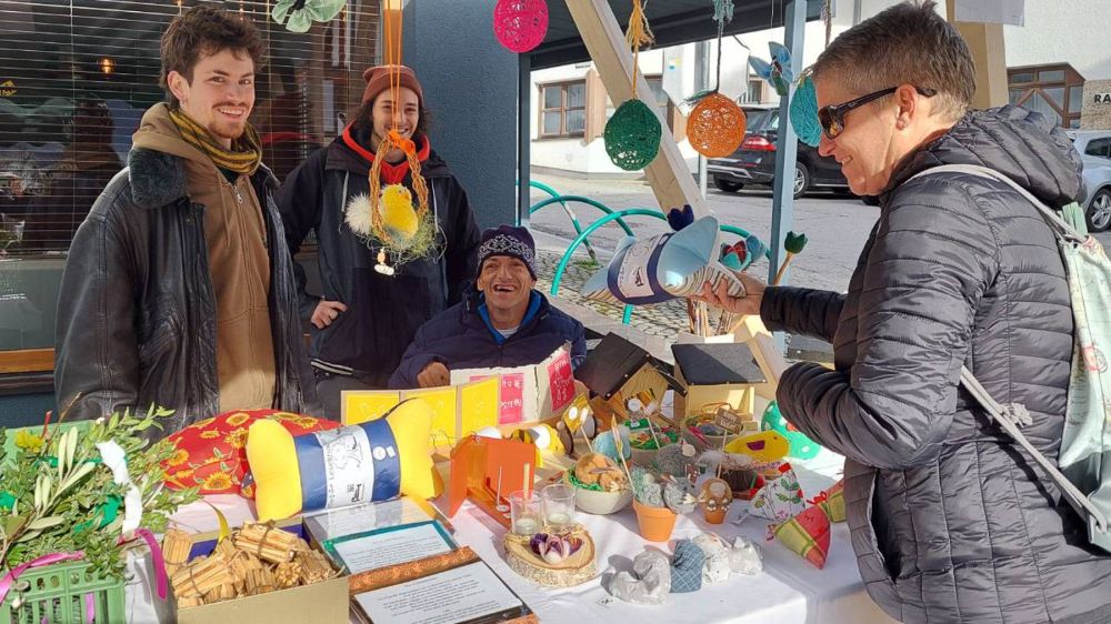 drei Männer bieten Waren an einem Marktstand an; eine Kundin mit Sonnenbrille sieht sich die Waren an