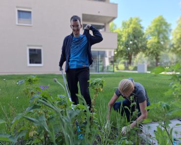 Ein Mann mit Behinderung und eine Unterstützerin arbeiten im Garten einer Wohnanlage an einem grünen Beet