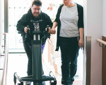 eine Frau mit Gehbehinderung übt mit einem Gehtrainer; die Unterstützerin geht neben ihr