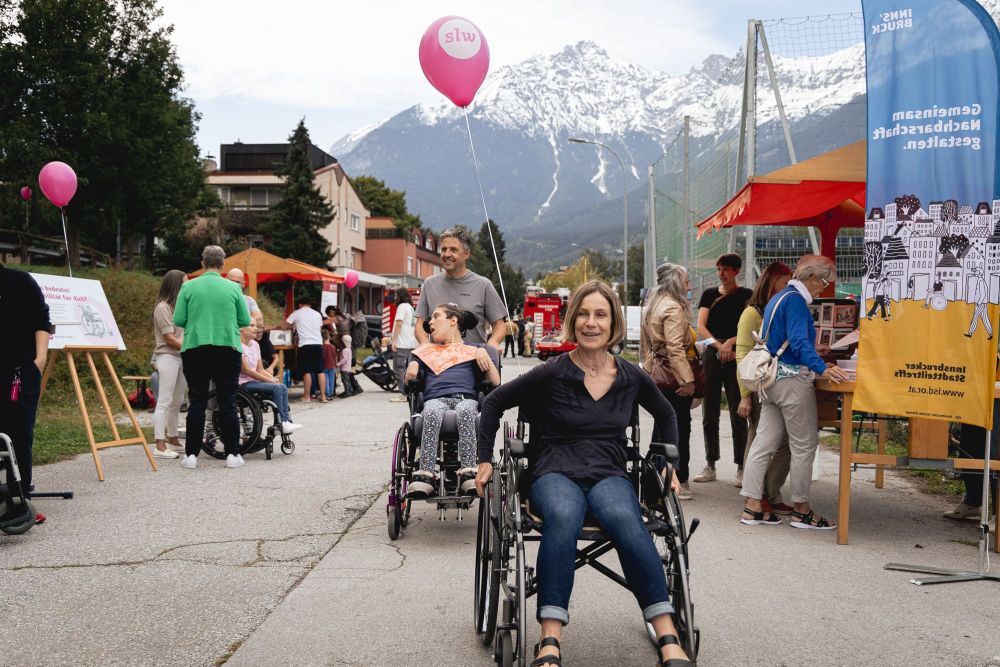 eine Frau im Rollstuhl lächelt in die Kamera; dahinter schiebt ein Mann eine weitere Frau im Rollstuhl; pinke Luftballons und bunte Straßenfest-Buden im Hintergrund