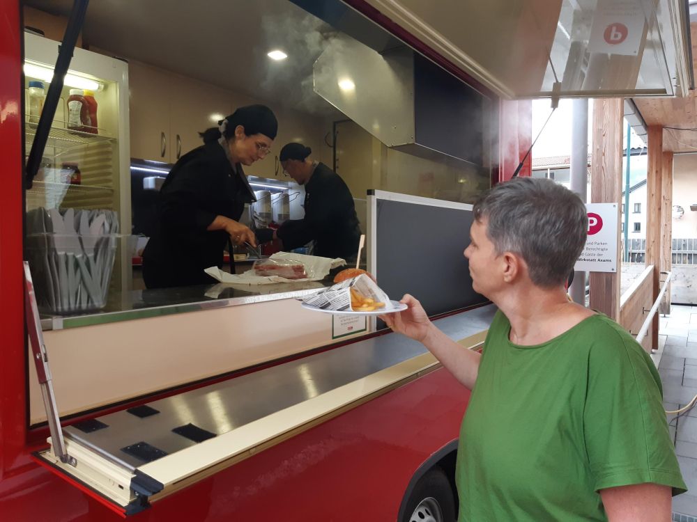 eine Frau erhält einen Teller mit Burger und Pommes aus einem roten Foodtruck; darin arbeiten eine Frau und ein Mann, die das Essen zubereiten