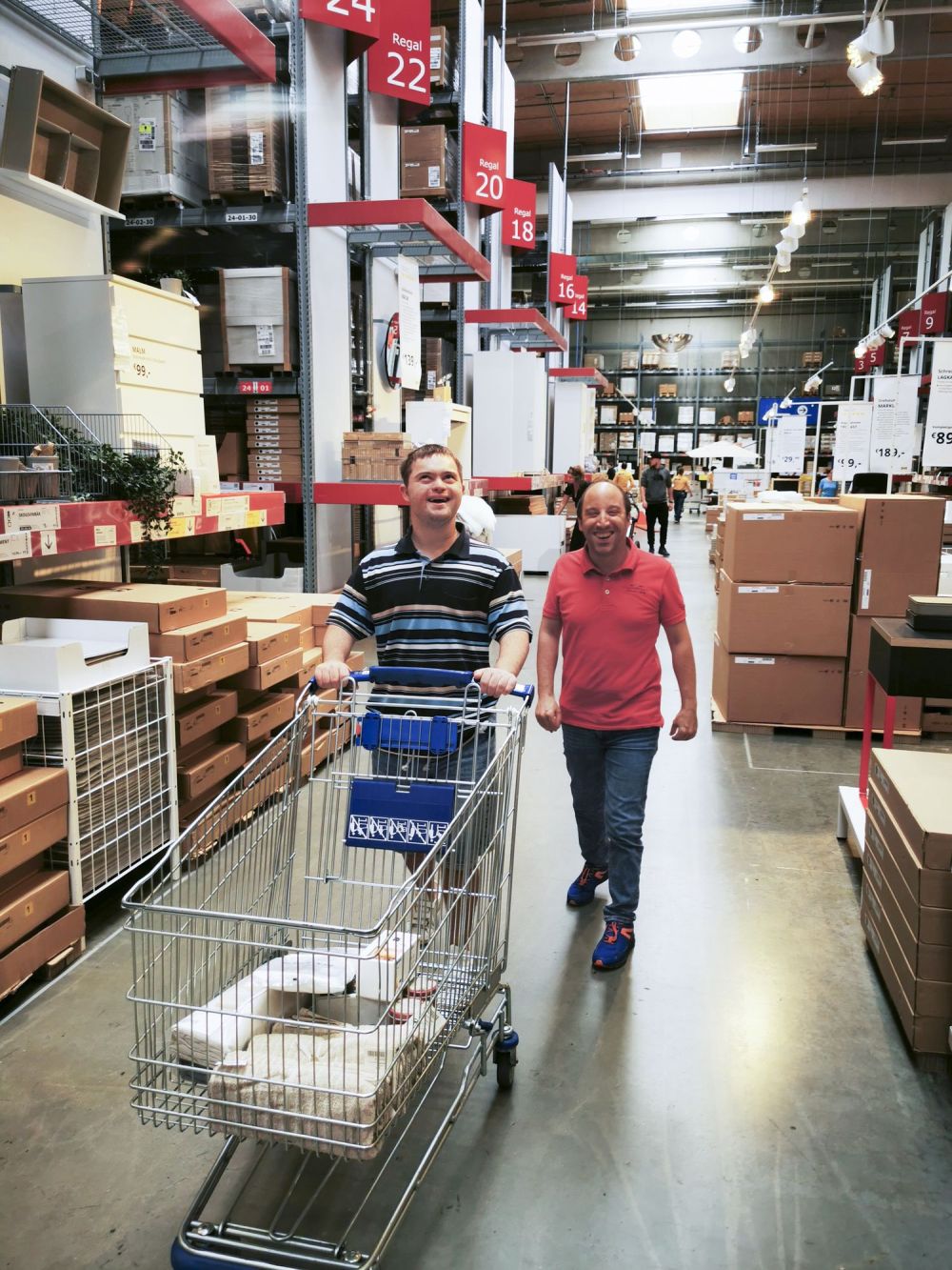 zwei Männer mit Behinderung schieben einen Einkaufswagen durch das Lager eines Möbelhauses