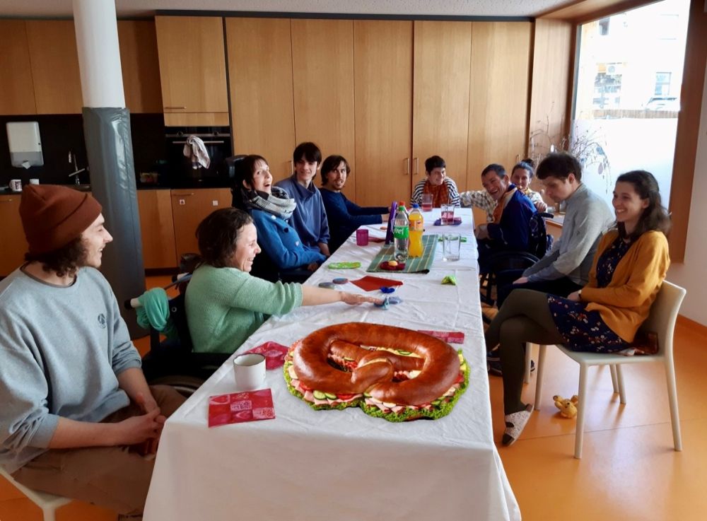 eine Gruppe von Menschen (teilweise mit Behinderungen) sitzen um einen langen Tisch mit Tischdecke; darauf ist eine große Partybreze, Servietten und Getränke