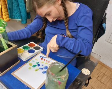 eine Frau im Rollstuhl zeichnet mit Fingerfarben auf einem Blatt Papier