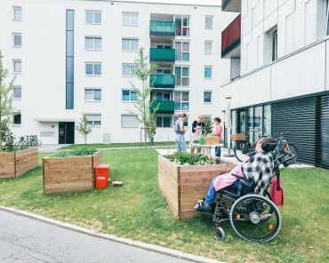 ein Garten in einer Hochhausanlage; mehrere Hochbeete; eine Person sitzt im Rollstuhl, einige weitere Personen stehen um die Hochbeete