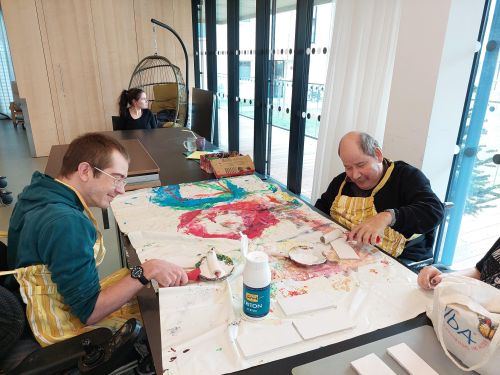 zwei Männer sitzen an einem großen Tisch gegenüber; mit Farbrollen rollen sie weiße Farbe auf kleine Holzstücke