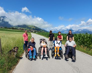eine Gruppe Menschen mit und ohne Behinderungen spazieren auf einem Feldweg an einem schönen, sonnigen Tag; Manche Menschen sitzen im Rollstuhl; links im Gras sitzt ein schwarz-brauner Hund; alle schauen freundlich in die Kamera